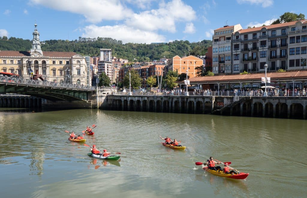 A group of kayakers on the river in Bilbao, kayaking underneath a stone bridge.