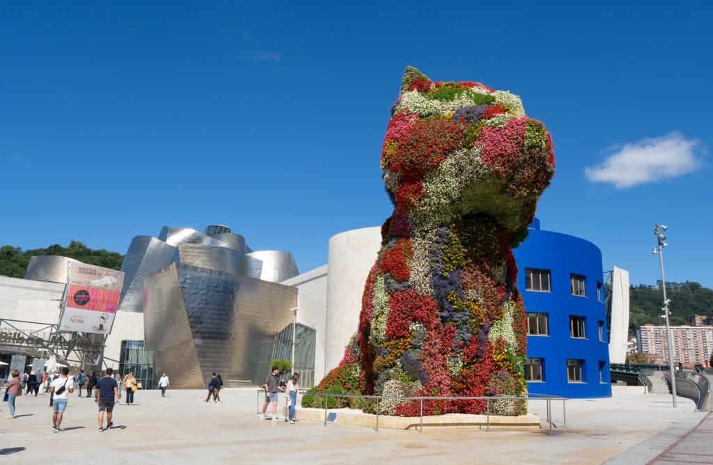 A giant flower-covered sculpture shaped like a terrier puppy in front of the shiny Guggenheim Museum.