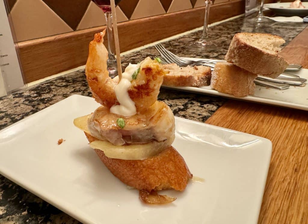 A Pintxos with a shrimp and some meat and cheese attached to a piece of bread with a toothpick.