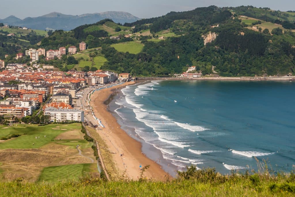 A small town of white buildings with orange roofs perched on the edge of the sea, with lots of big waves, and big green hills in the background.