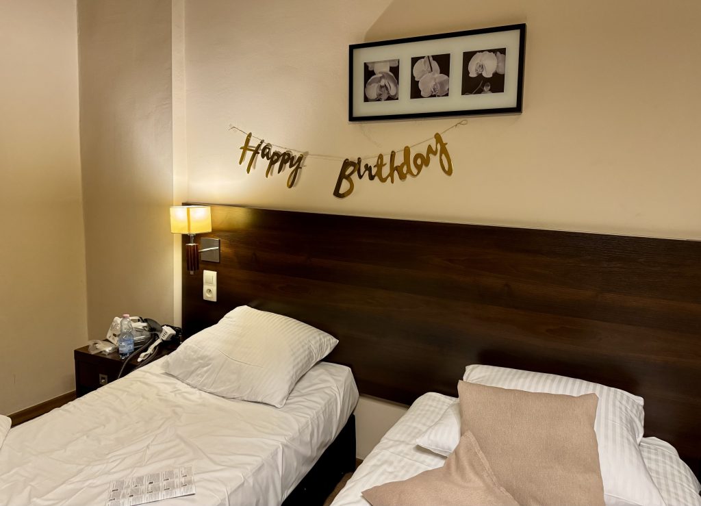 A hotel room with two twin beds and a gold "Happy birthday" banner hanging over one bed.