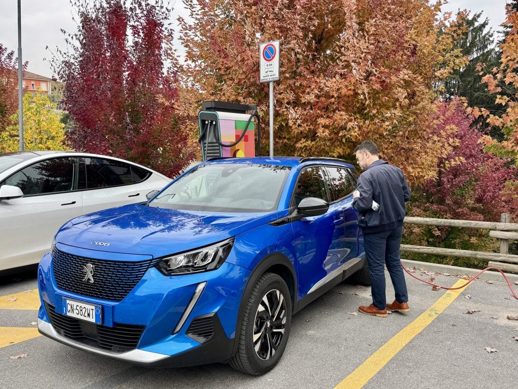 Charlie attempting to fuel up a bright blue electric SUV, surrounded by colorful fall foliage.