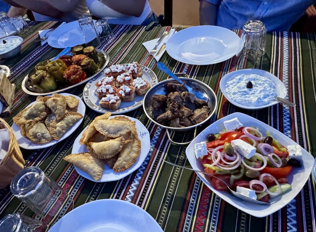Plates of Cretan dishes: fried empanada-like dumplings, roasted lamb, bruschetta topped with tomatoes and feta, Greek salad.