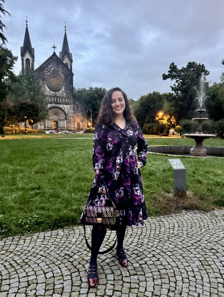 Kate standing in front of a church in Prague wearing a long raincoat with a full skirt in a floral pattern of black, deep purple, and lavender. She carries a purple and gold purse and wears burgundy patent leather heels and navy blue tights.