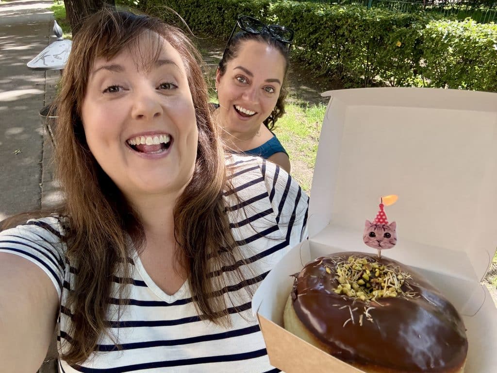 Kate and Cailin taking a selfie. Cailin holds a box with a Dubai chocolate donut with a lit candle shaped like a cat in a party hat sticking into it.
