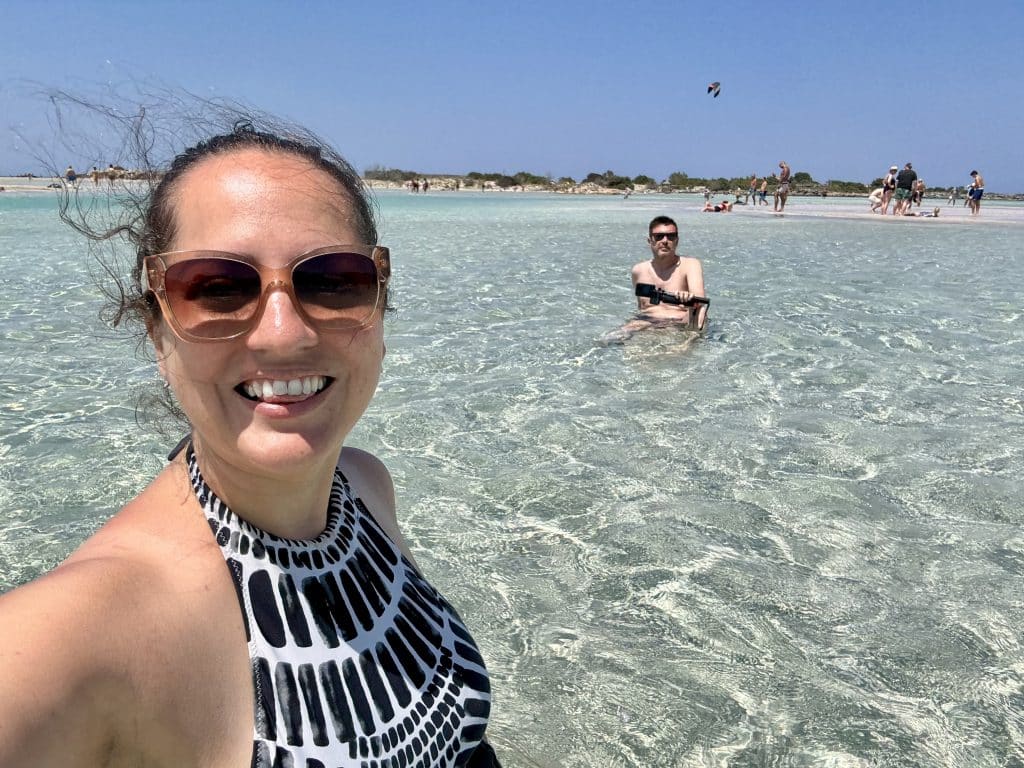 Kate and Charlie in the pale turquoise waters of the sea in Crete, Kate taking a selfie and Charlie far in the background.