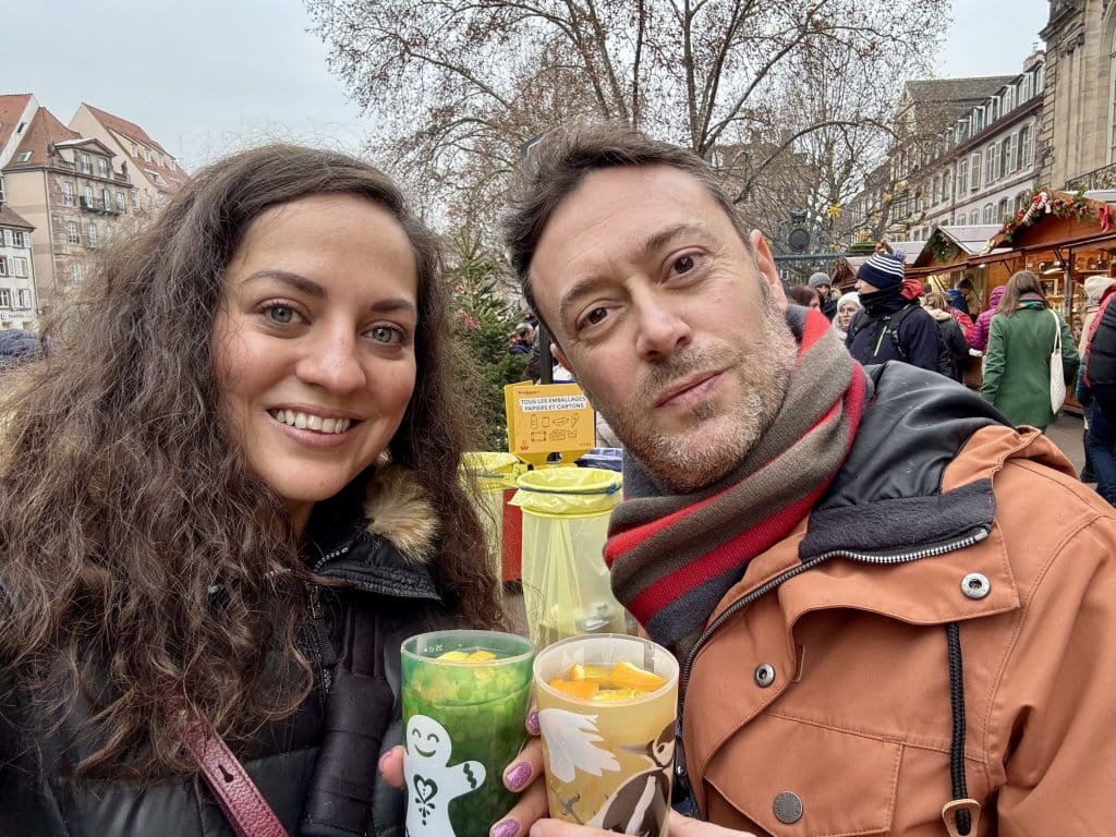 Kate and Charlie smiling, holding plastic cups filled with white mulled wine and slices of fruit.