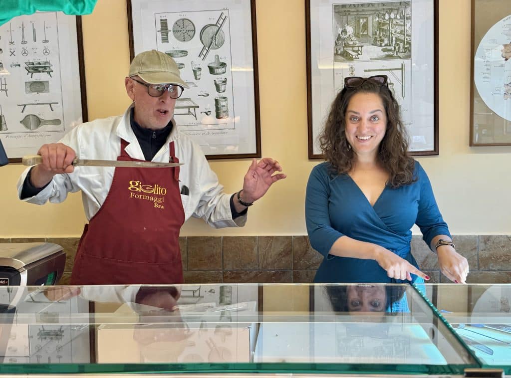 Kate standing in a cheese shop behind a counter. Next to her is a man in an apron holding a knife and pointing it at her like a villain.