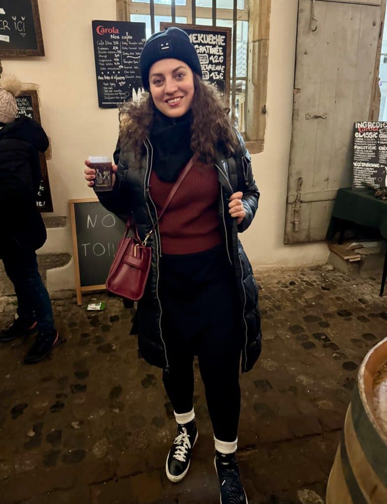 Kate wearing a winter coat on top of a sweater over a dress, leggings, wool socks, and sneakers, plus a beanie on her head.