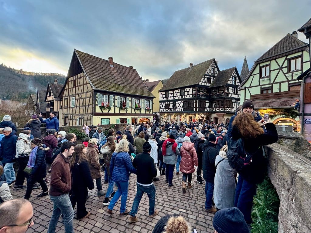 Huge crowds of people on a square in France, surrounded by fairy tale half-timbered houses with thatched roofs.
