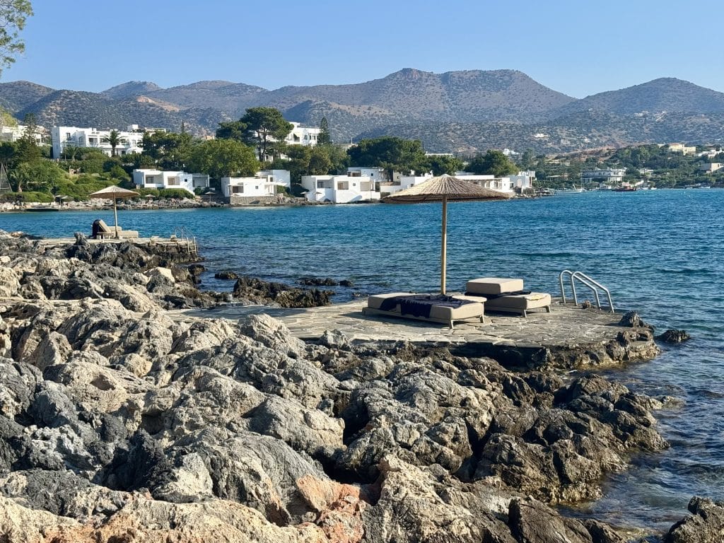 A rocky beach set on bright blue water, with a few beach chairs beneath palapas.