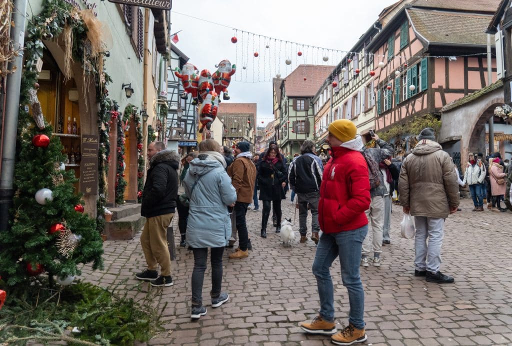 A cobblestone street full of people, surrounded by peach and green half-timbered houses, Christmas lights running between the two sides of the street.