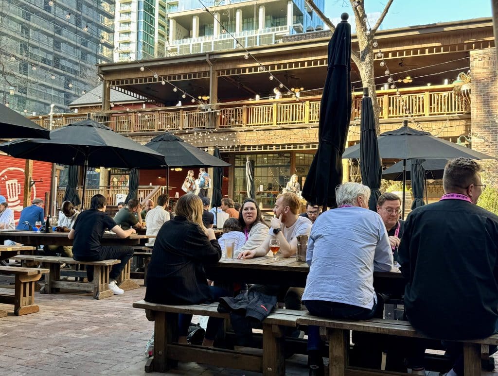 People sitting outside an old-fashioned wooden bungalow turned into a bar, with high-rise condos in the background.