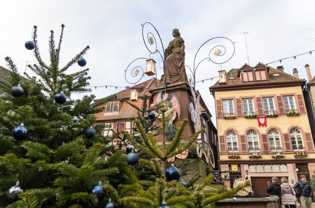 Christmas trees topped with dark blue ornaments in the foreground, and a statue and peach-colored houses in the background.