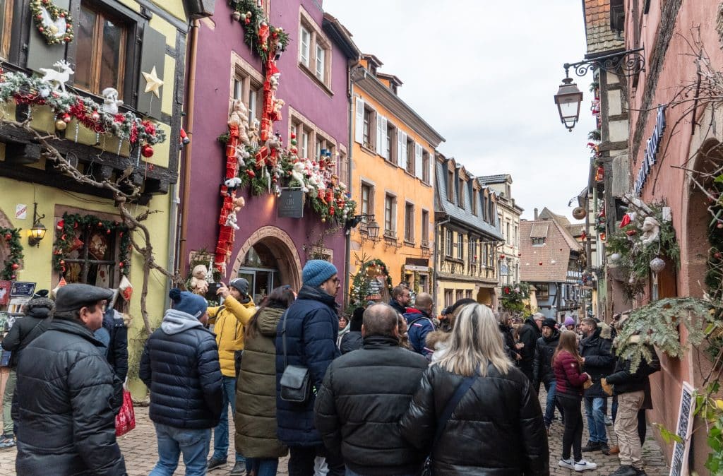 Big crowds of people walking down a narrow street filled with colorful half-timbered homes covered with Christmas garlands.