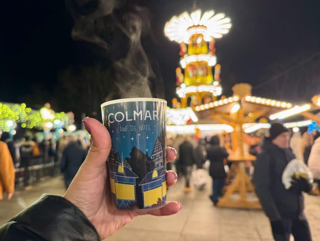 A steaming plastic cup of hot wine that says Colmar on it, and the market in the background