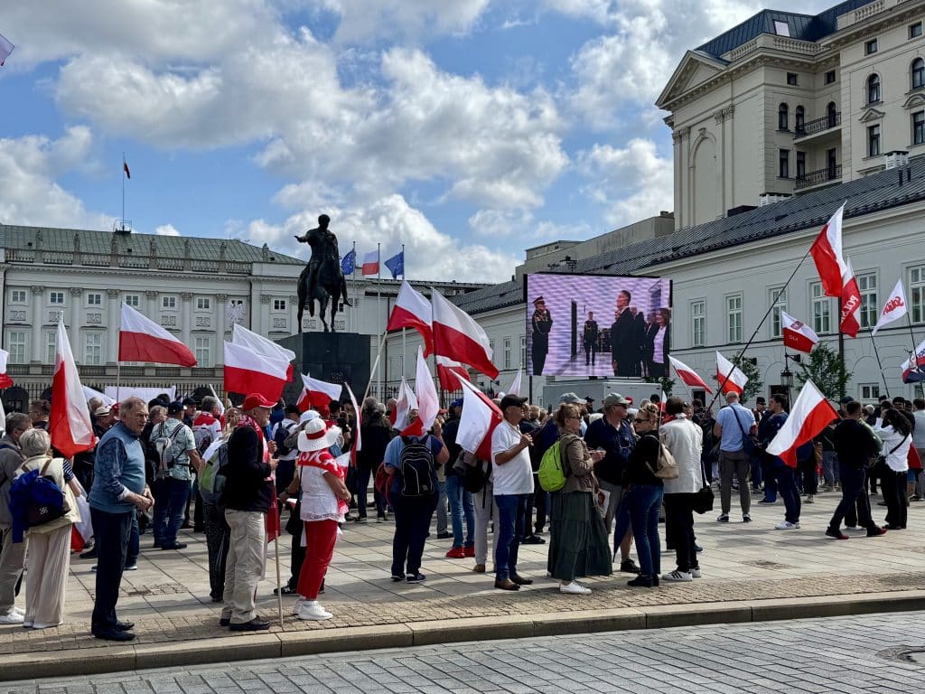 A group of people holding Polish flags outside the Warsaw presidential building as the new president takes the oath on screen.