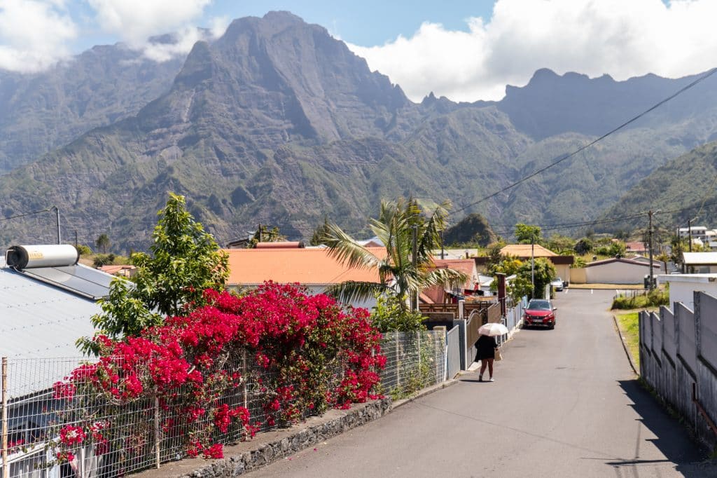 A woman in an umbrella walking down a calm street filled with cottages and lots of flowers. Mountains are in the background.