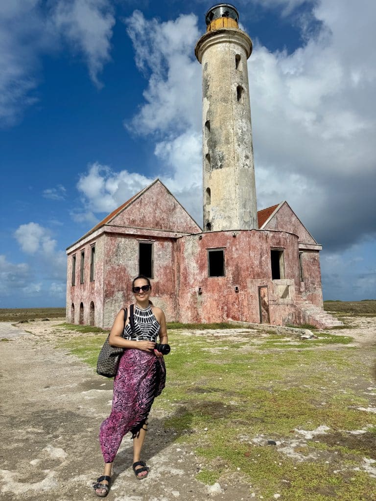 Kate in a bathing suit and sarong standing in front of a pink and white lighthouse on an island.