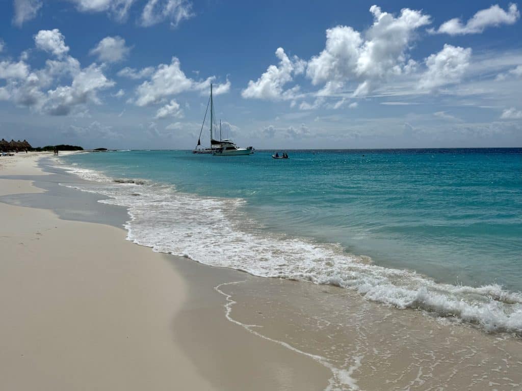 A white sand beach with bright turquoise water, softly crashing waves, and a sailboat in the distance.