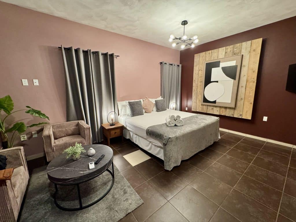 A calm and tastefully decorated hotel with pink walls and white and gray bed with gray curtains and a tan chair.