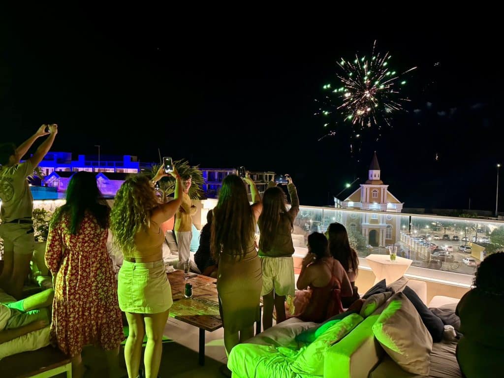 A group of women standing on a rooftop at night, looking at fireworks over a church.