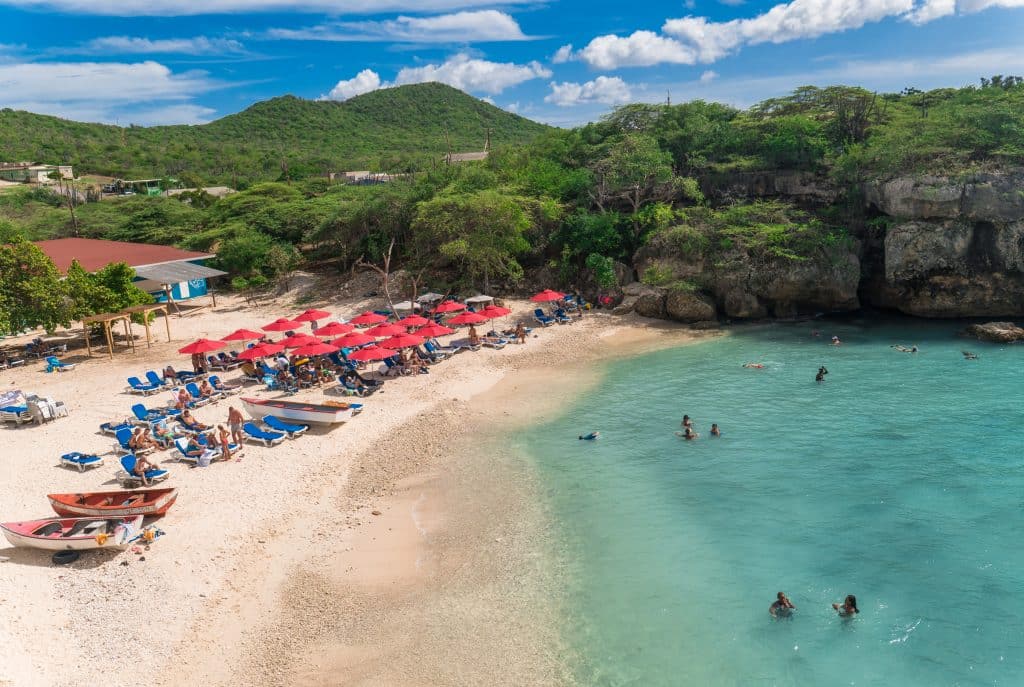 A beach with red umbrellas over beach chairs, and bright teal calm water.