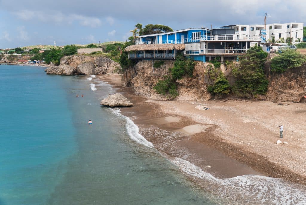 A small beach next to a cliff with houses on it, perched above a bright turquoise sea.
