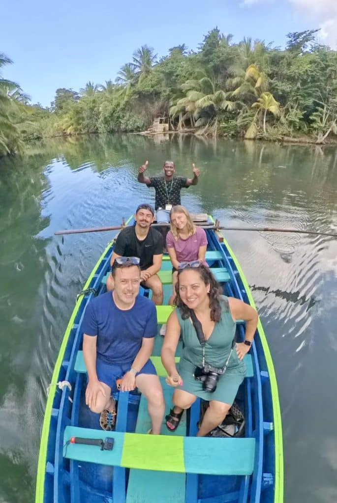 Kate, Charlie, and two other people with a driver in a wooden rowboat, traveling along a calm river surrounded by palm trees.