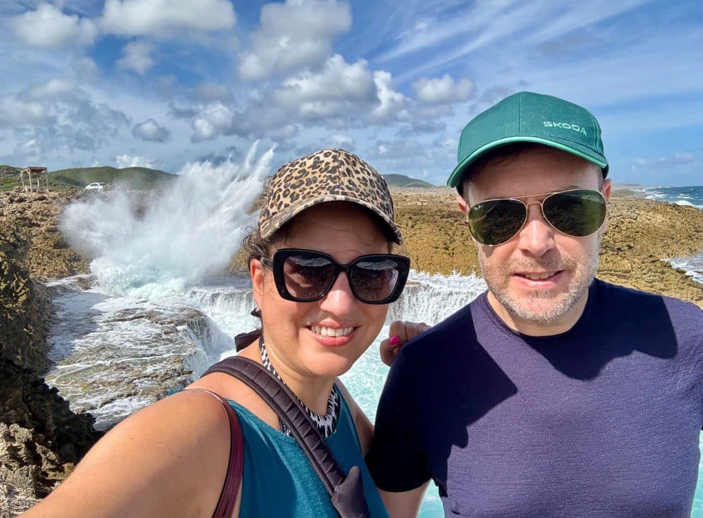 Kate and Charlie wearing baseball caps and standing in front of a giant crashing wave.