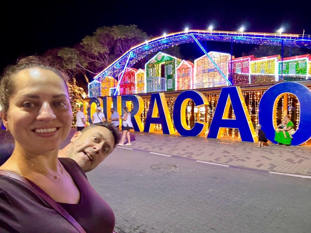 Kate stands in front of big Curacao sign at night, while Charlie pops out behind her and looks like a tiny head sticking out of her shoulder.