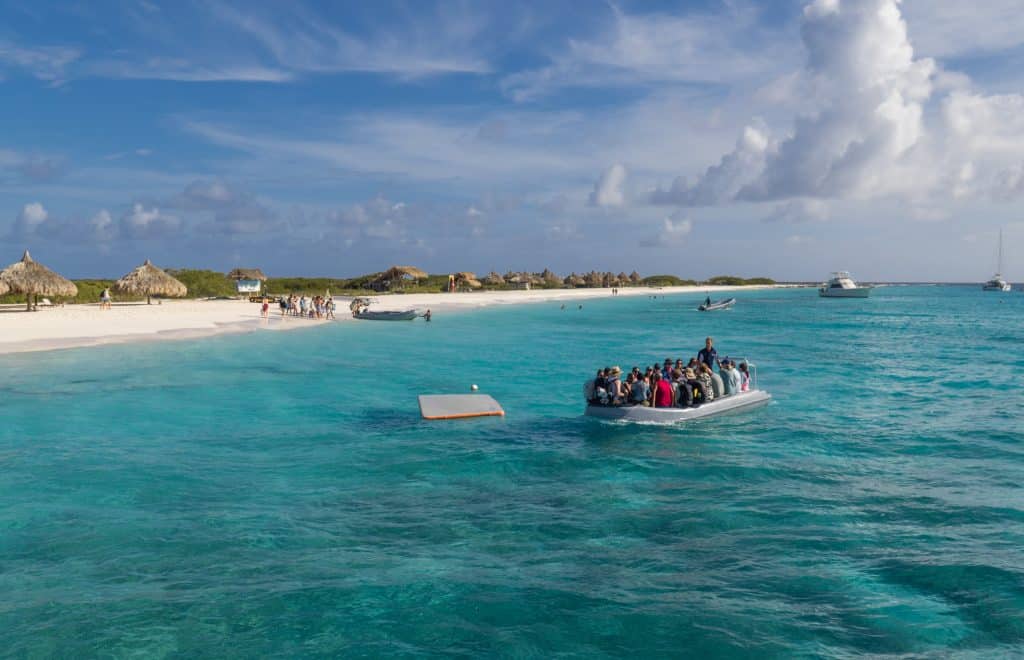 A rubber zodiac boat transferring people back to a boat in the middle of bright blue water, just off a white sand beach.