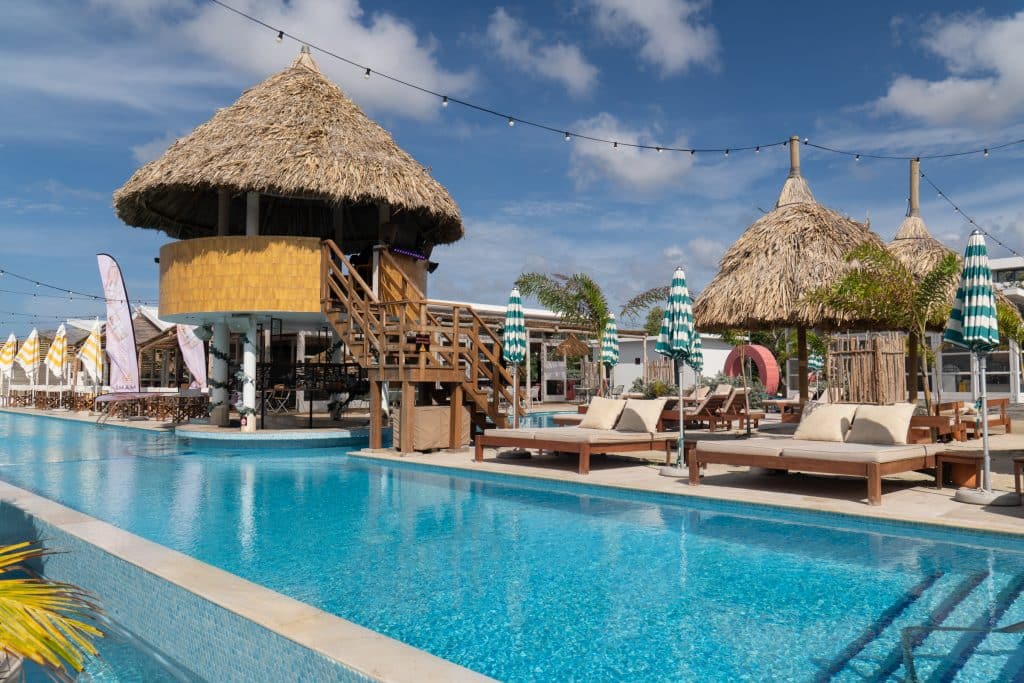 A beach club with a calm long pool surrounded by beach chairs and palaces.