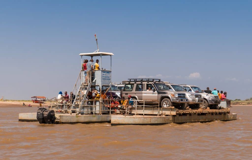 A larger raft perched on canoes with several SUVs and tourists making the journey across a brown river.