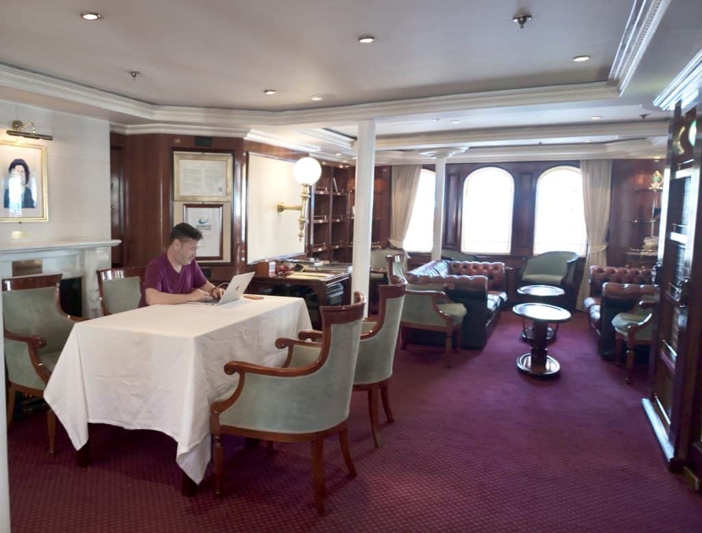 Charlie sitting at a table and working on his laptop in a small library with bookshelves and big windows overlooking the sea.