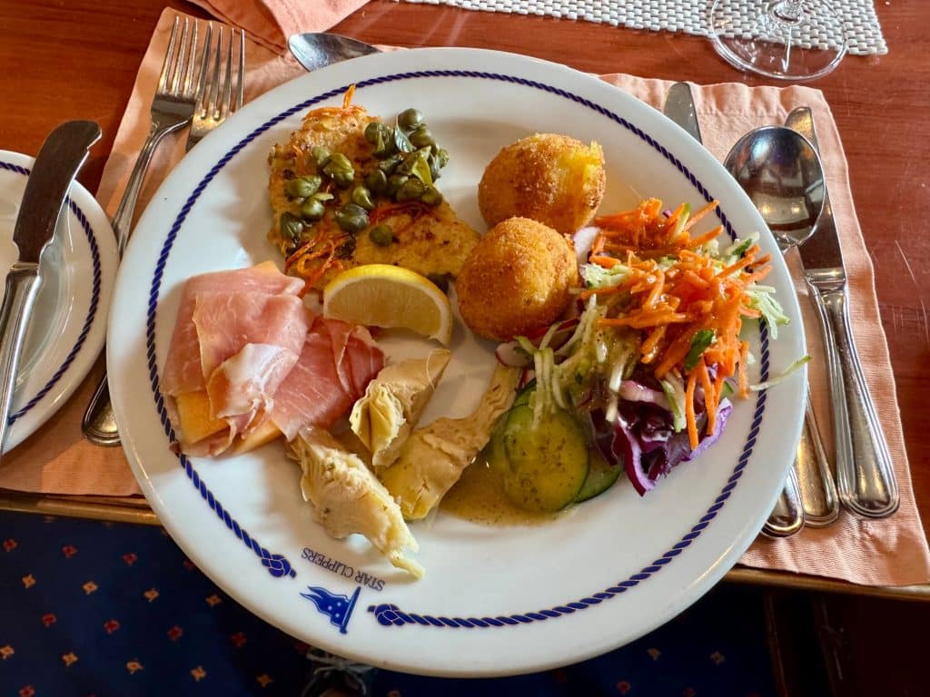 A buffet plate filled with chicken piccata, arancini, prosciutto and cantaloupe, and salad.