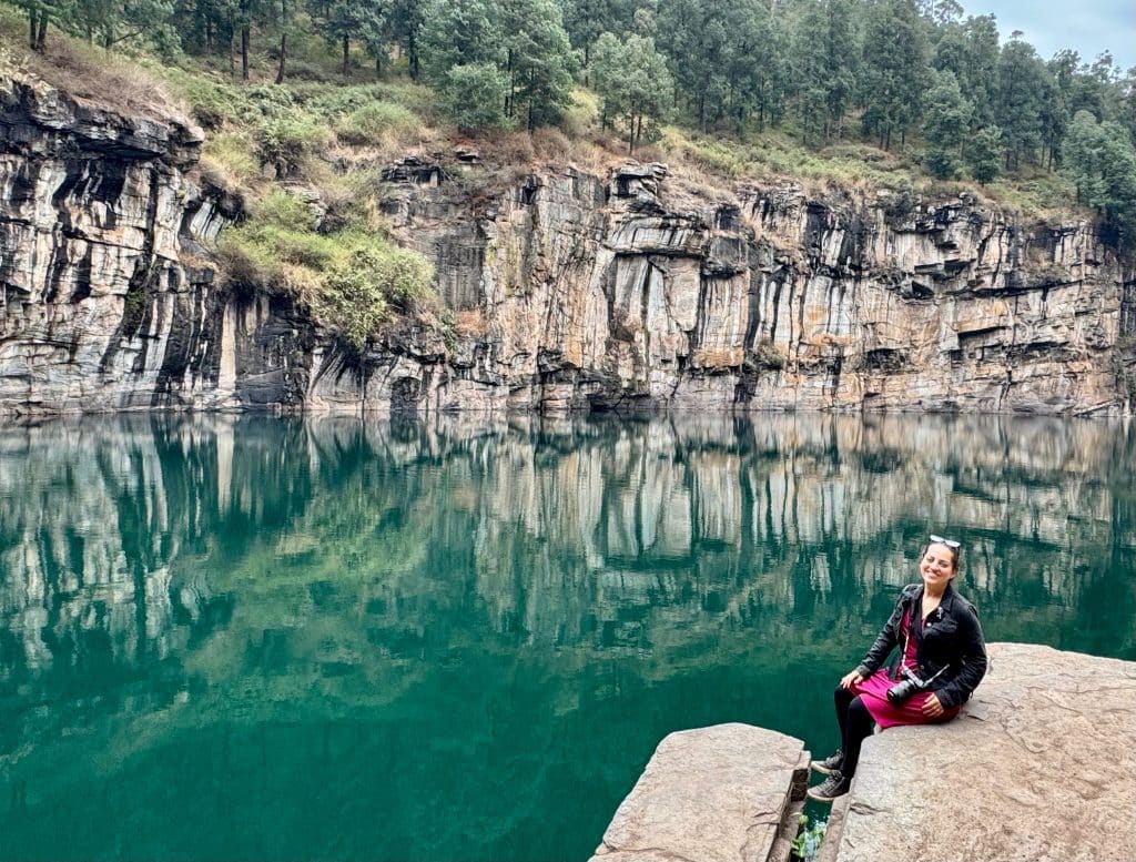 Kate wearing a pink dress and black jacket and sitting on the edge of a deep teal lake surrounded by stone edges.