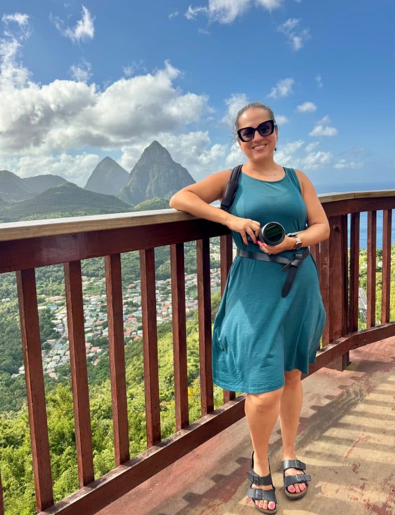 Kate wearing a sleeveless teal dress and holding a camera while standing in front of a fence overlooking the conical mountains of St. Lucia.