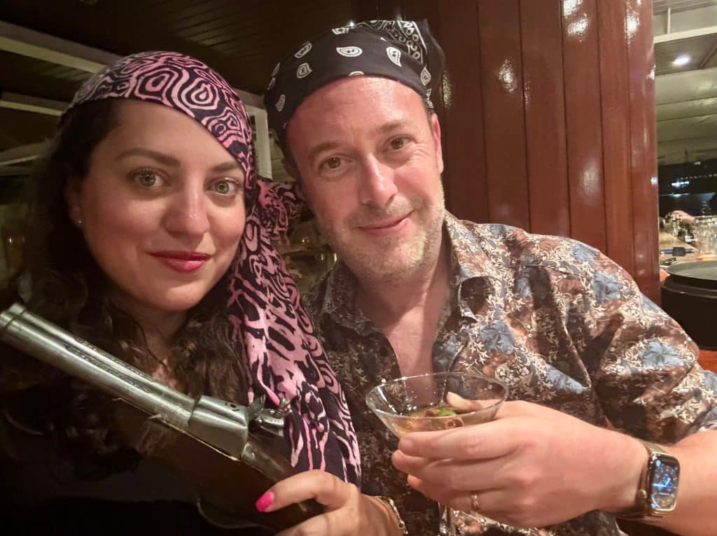 Kate wearing a pink and black scarf soon her head, Charlie wearing a black bandana on his head. Kate holds a fake pistol and Charlie holds a martini.