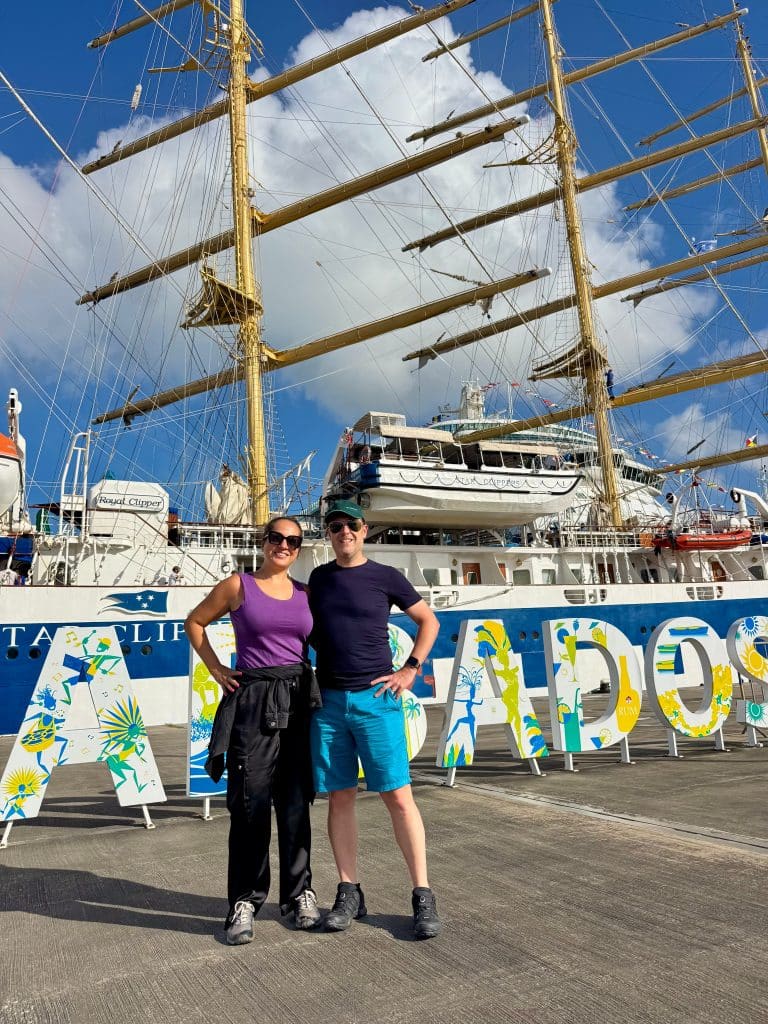 Kate and Charlie standing in front of the Barbados sign with the masts of the Royal Clipper behind them.