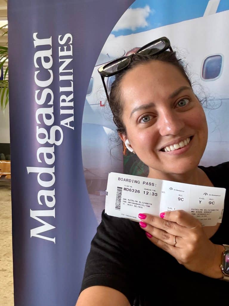 Kate standing in front of a Madagascar Airlines banner and holding a ticket from Antananarivo to Nosy Be.
