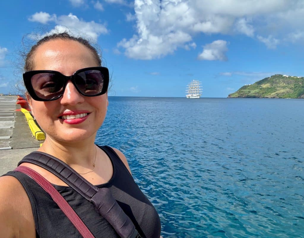 Kate taking a smiling selfie in black sunglasses. Behind her in the far distance is the Royal Clipper anchored in the sea, its five masts towering high.