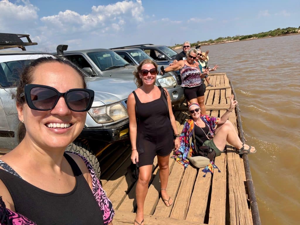 Kate and friends standing on a wooden raft crossing a brown river with SUVs parked behind them.