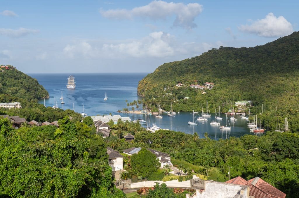 A gorgeous little bay in the Caribbean filled with lots of sailboats, and the Royal Clipper tall ship in the distance, It's surrounded by lush green vegetation on all sides.