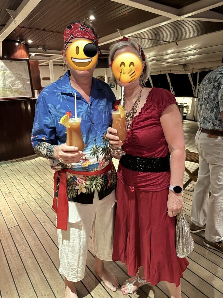 A middle-aged couple (faces obscured with emojis) dressed up in full pirate regalia and holding tropical drinks.