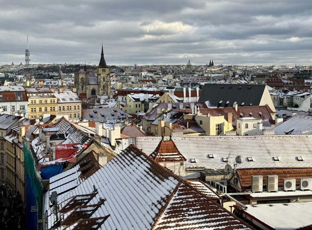A roof of snowy rooftops in Prague, with lots of church steeples and orange roofs pointing up.