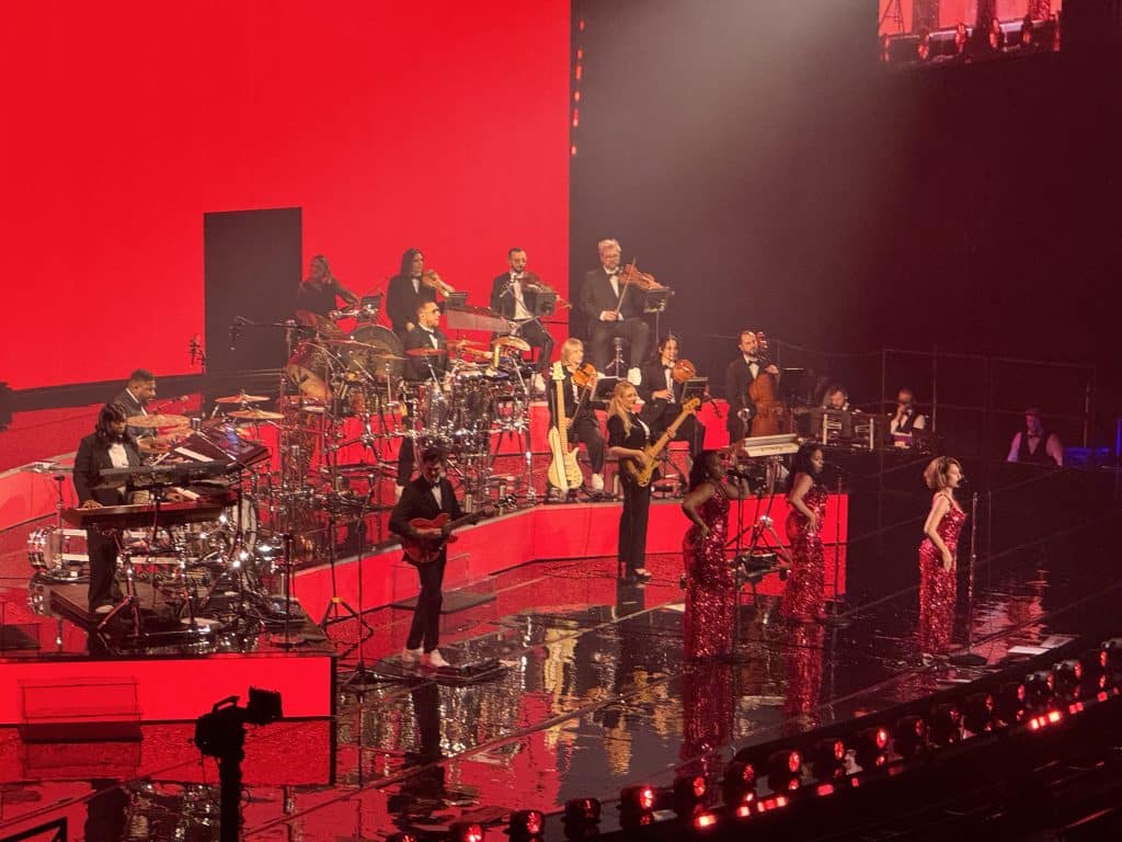 Singer Raye performing in a long red sequined gown with a 1940s-style big band behind her.