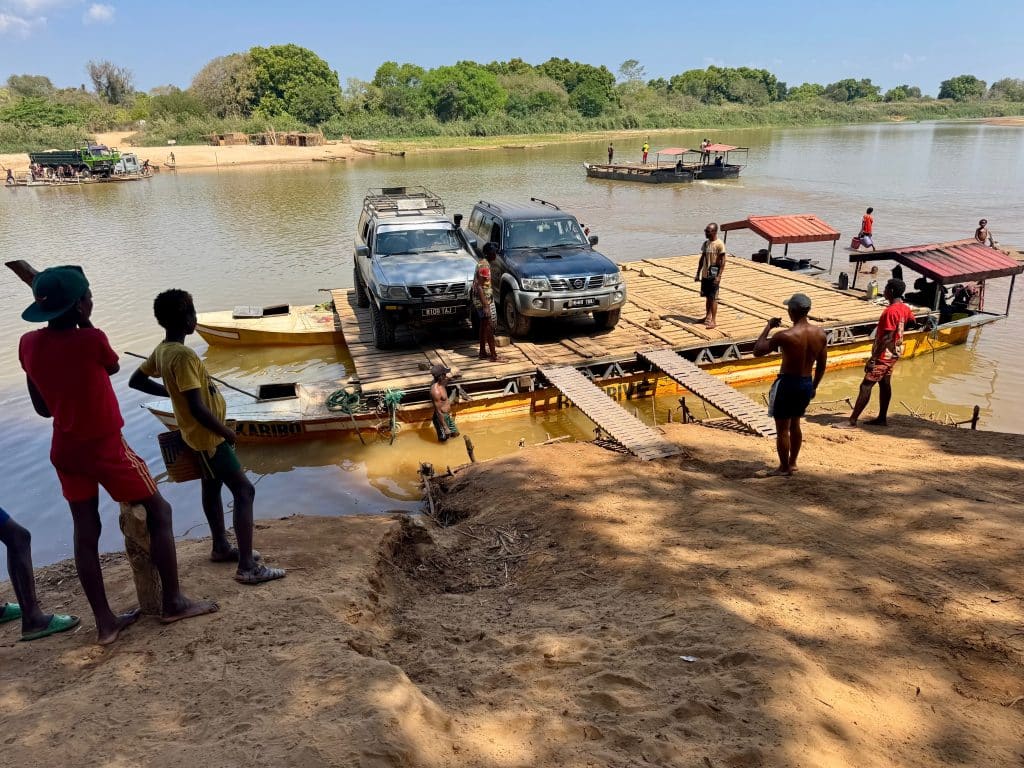 A few SUVs parked on a primitive looking river raft with planks perched on top of canoes, as local men look on.
