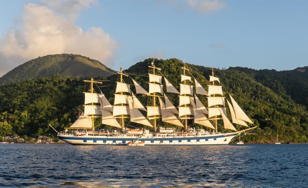 A tall ship sailing with so many sails, illuminated by the sun, tall green mountains in the background.