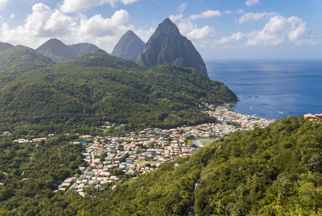 A few of a small city on the edge of the sea, surrounded by mountains covered with greenery and the two green pointy Piton mountains in the distance.
